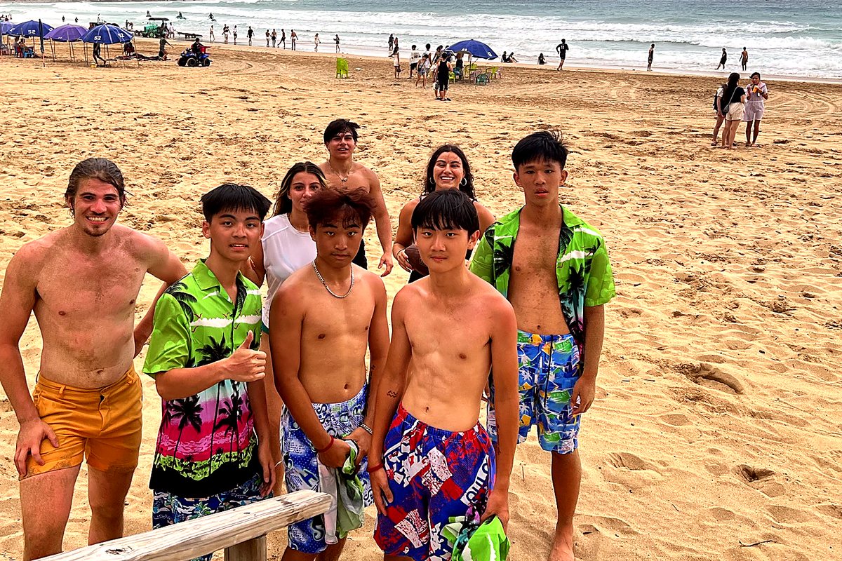 Eight young people standing together on the beach in Taiwan, several in matching green palm-print shirts, ocean behind them.